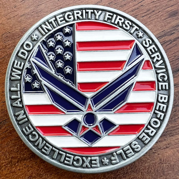 Round silver challenge coin with U.S. flag centered in background. USAF eagle logo atop flag. Text around image. "We do integrity first service befor self excellence in all"