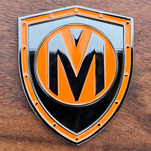 A custom shield-shaped challenge coin with an orange "V" and black "M" logo. 