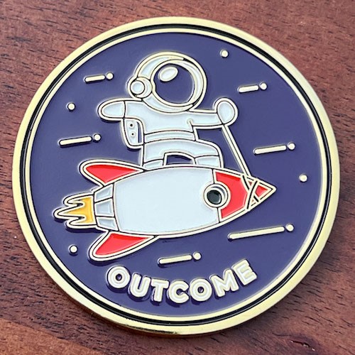 Round coin, silver base. Astronaut standing on lassoed rocket on dark blue background. White stars in backgroun. Rocket finished in red and yellow